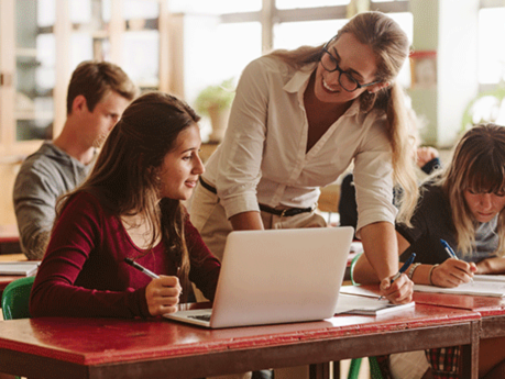 Female teacher with long blonde hair and glasses leaning on student desk helping with her work.